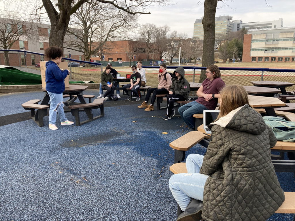 Eight students are outside next to the Honors House at the tables. One student stands near a table with a cardboard box on top of it and presents something to the other seven students. There are a few trees in the background, all without leaves.