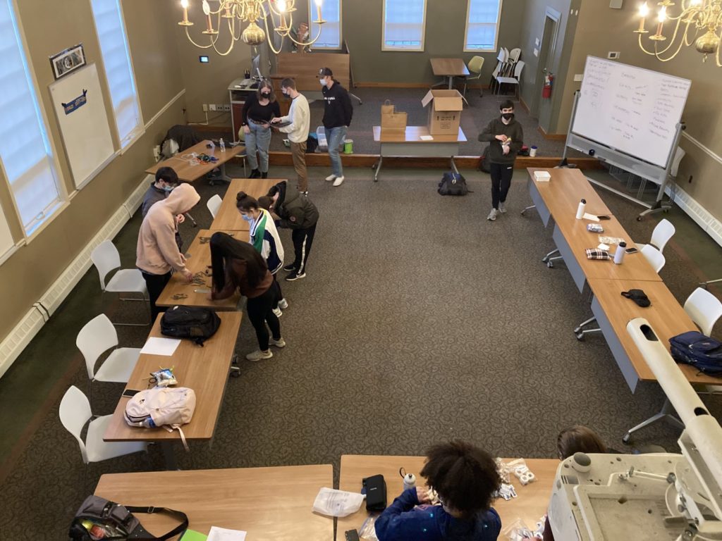 Many long, wooden tables form a horseshoe in the center of the Honors House Lyceum. Students stand around the tables and work on projects.