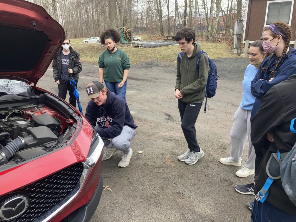 Seven students stand around a car with an open hood. One student crouches down near the left front tire and appears to be explaining something to the students standing around them in a half circle. There are many skinny trees in the background without leaves.