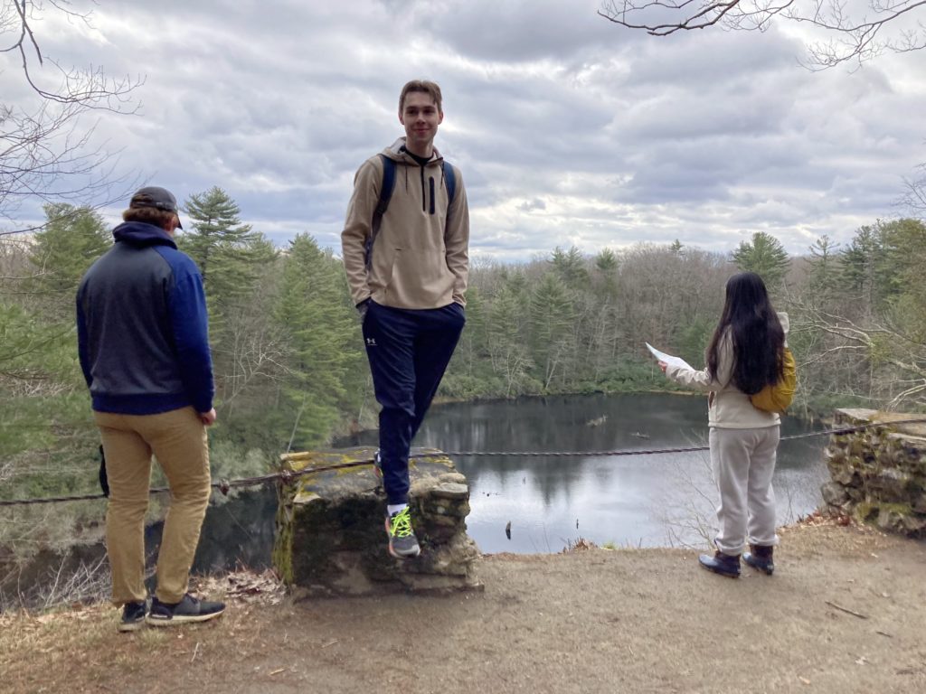 Three students stand near the edge of a pathway overlooking a large lake. The lake is surrounded by evergreen trees. Two students look at the lake. The student on the right holds a piece of paper. The student in the middle is stepping off a pile of rocks near the edge.