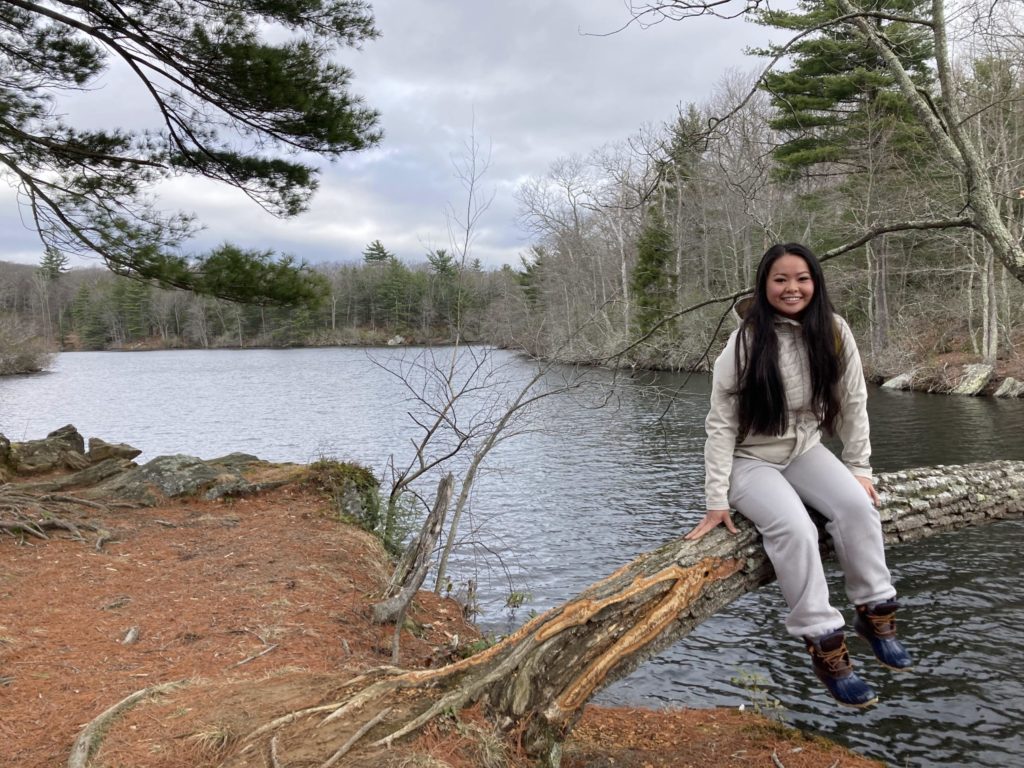 Student sits on a tree trunk that is almost coming out of the ground. The bottom of the trunk is somewhat stripped of bark. There is a huge lake in the background with many trees without leaves and some pine trees surrounding it.