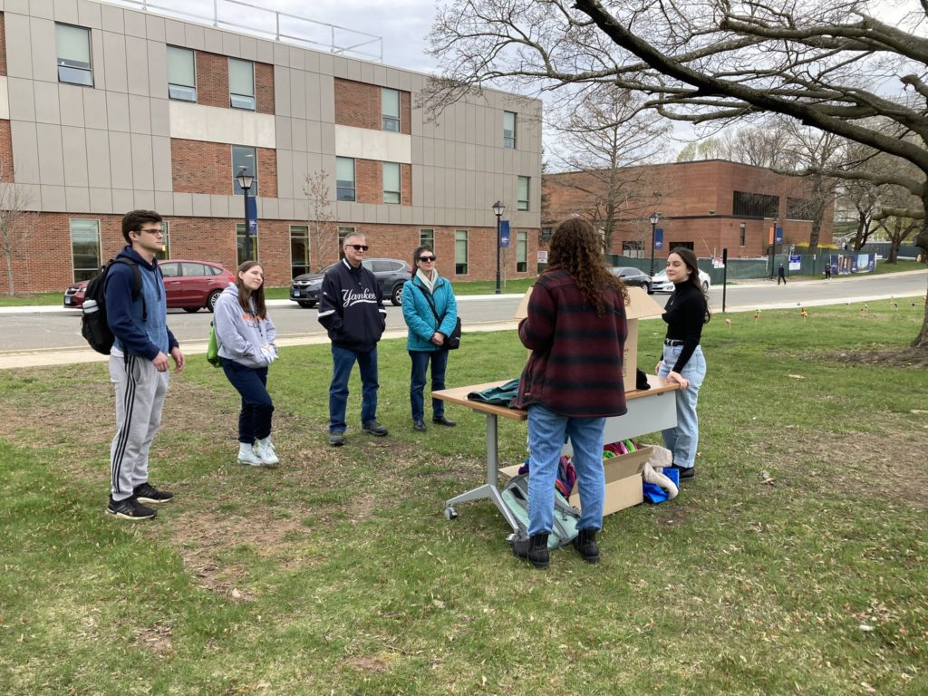 Two students present a project to four visitors outside the Higgins Building. The students are standing on either side of a wooden table, and there is a large cardboard box between them on the table.