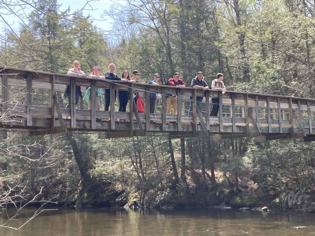 Nine students standing on a long wooden bridge above a big river. There are many trees around them, some without leaves and some evergreen.