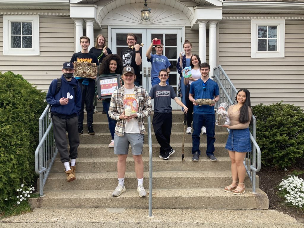 Twelve students stand in front of the honors building holding up projects they worked on that represent who they are. They include clay sculptures, walking sticks, and multimedia 3D art. There are large green bushes and small white flowers on either side of the steps.