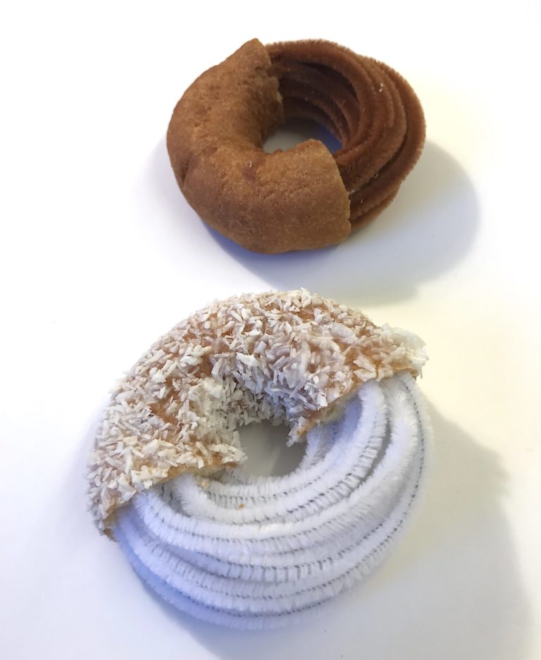 Two inedible donuts. The donut in the front is made of pipe cleaners and white flakes, creating a glazed appearance. The donut behind it is made of brown pipe cleaners and some kind of felt.