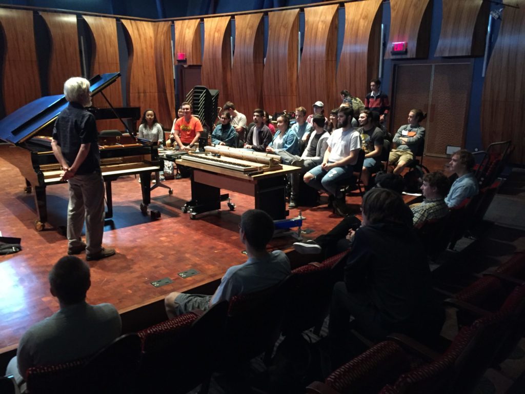 Students sitting stage left listening to Chris Farrell giving a presentation. Farrell is in front of a keyboard, and there is a grand piano to his left. A few people sit in the front row of the theater's orchestra section.