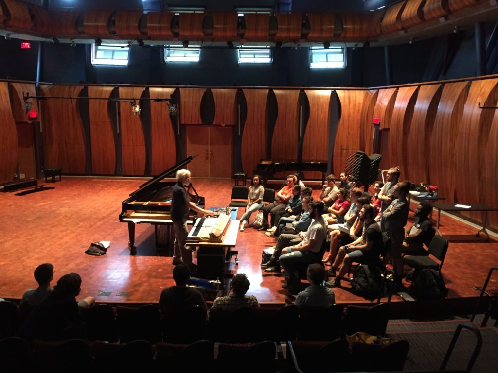 Students sitting stage left listening to Chris Farrell giving a presentation. Farrell is in front of a keyboard, and there is a grand piano to his left.