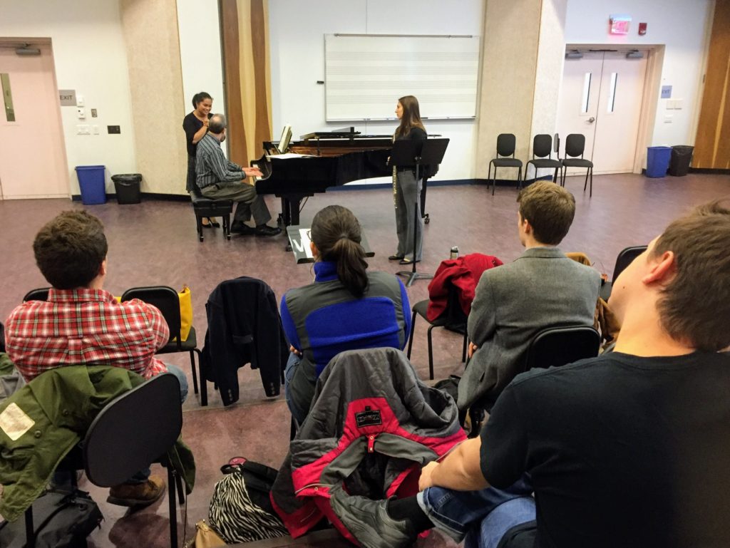 Two students at grand piano with Dr. Uriel Tsachor. Other students watch the masterclass from the other side of the room.