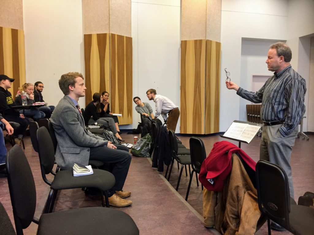 Dr. Uriel Tsachor, glasses in hand, talks to a student during the presentation. Other students sit and discuss on the other side of the room