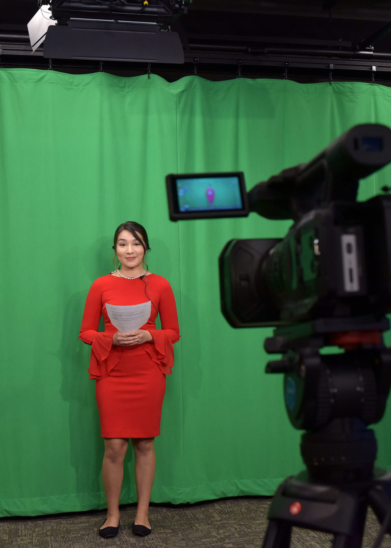 Female-presenting news anchor standing in front of green screen curtain. The student is wearing a dark orange dress and black flats. There is a camera in the foreground on the right recording them.