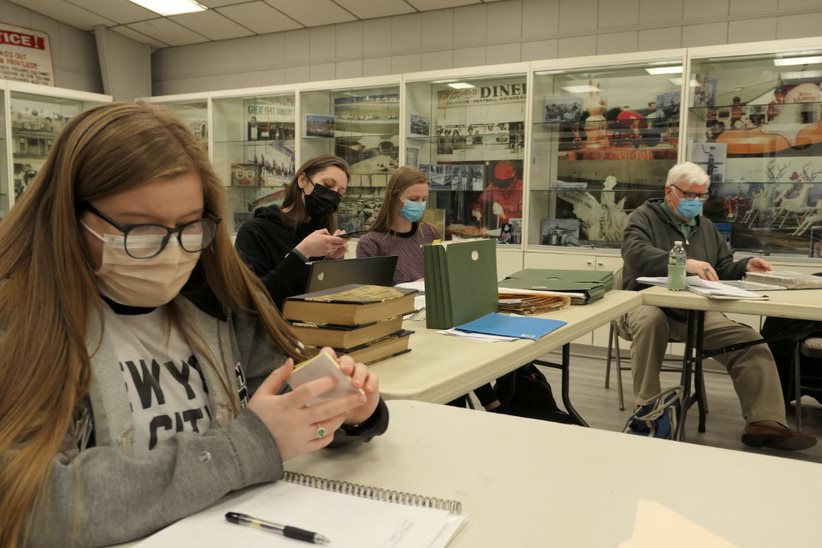 Students sit at tables taking notes on paper and laptops. They look at old, yellowing books and files. There are shelves behind them with photos from historical Danbury.
