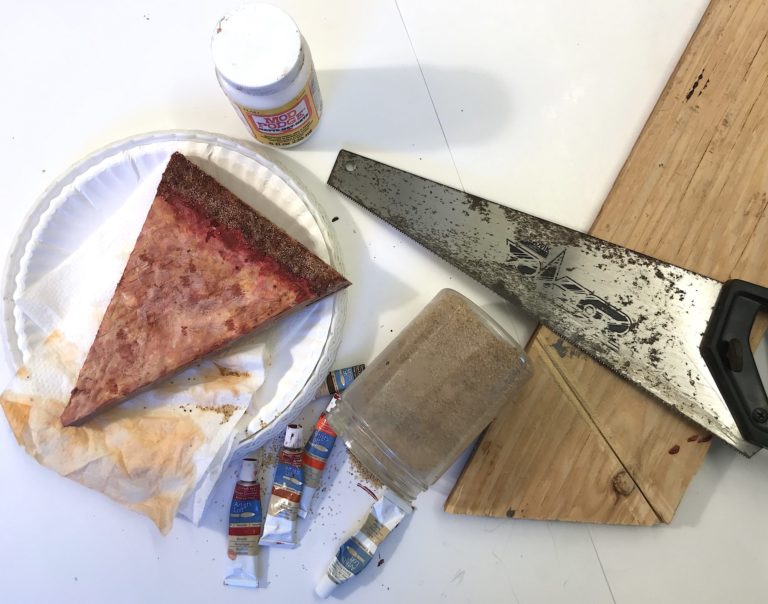 A pizza slice made of wood is on top of a stack of napkins and paper plates. Next to the slice are paint tubes, a jar of Mod Podge, wood shavings, a saw, and a plank of wood.