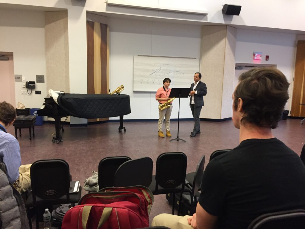 Saxophonist Dr. Kenneth Tse conducts a masterclass with a student playing a saxophone. Students watch from their seats on the opposite side of the room.