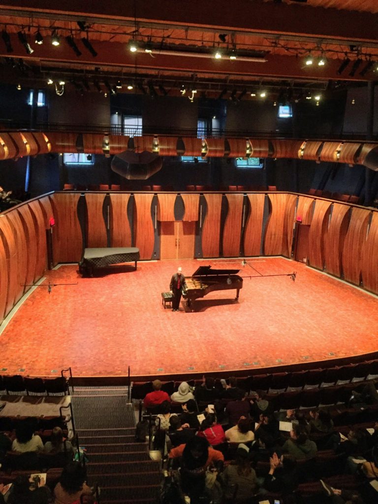 Dr. Uriel Tsachor stands next to a grand piano in the center of a large stage.