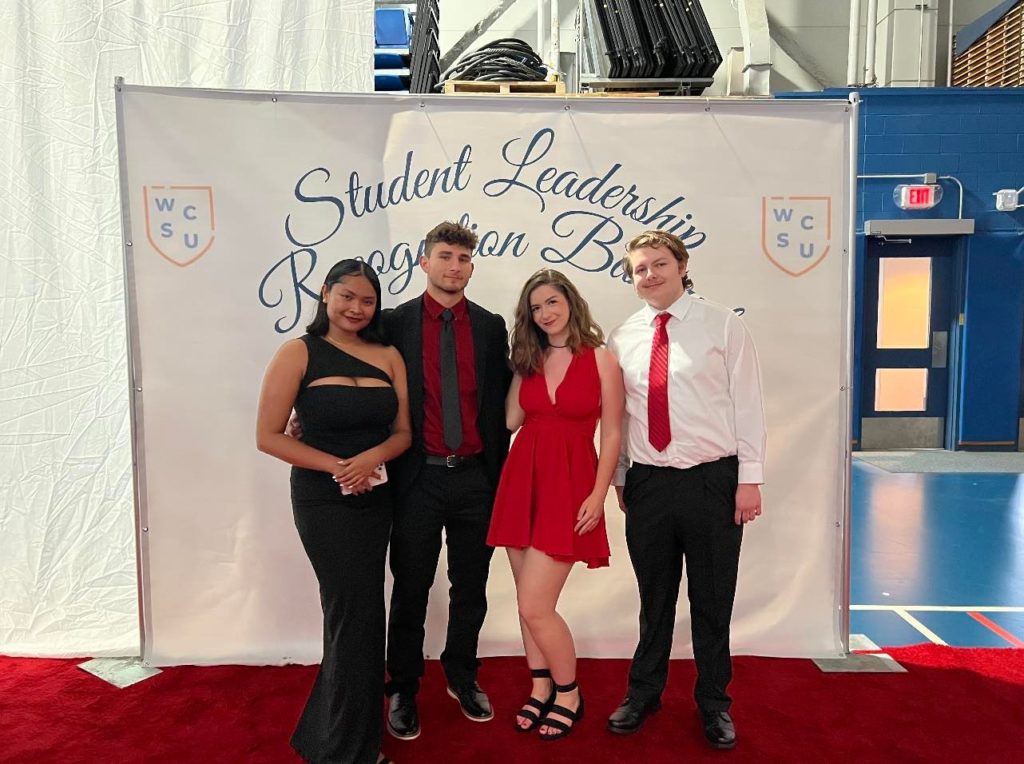 Four members of the HSOC E-Board stand in front of a Student Leadership Recognition Banquet backdrop. They are wearing black and red formalwear.