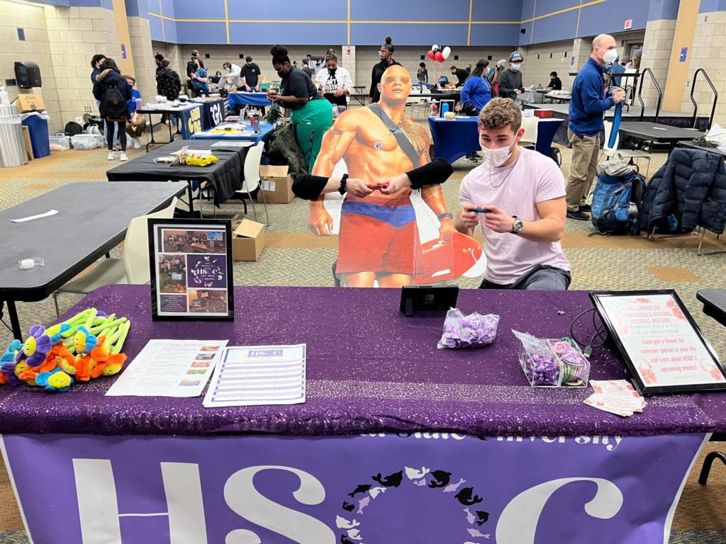 Members of HSOC E-board at the Clubs Carnival in the Westside Ballroom at the Campus Center. One member is pretending to battle a cardboard cutout of Dwayne "The Rock" Johnson in a game on the Nintendo Switch. Another member has their arms with a controller wrapped around the Rock to make it look like he's playing the game. The HSOC table is very purple and a poster with the logo, complete with an O made of many small dolphins, hanging off the front.
