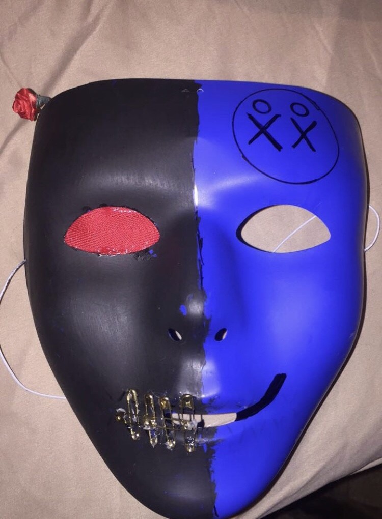A plastic blue mask painted black on the left side. On the right near the forehead, there are two stick figure drawings circled. There are safety pins across the left side of the mouth, and a piece of red ribbon taped inside the mask covers the left eye.