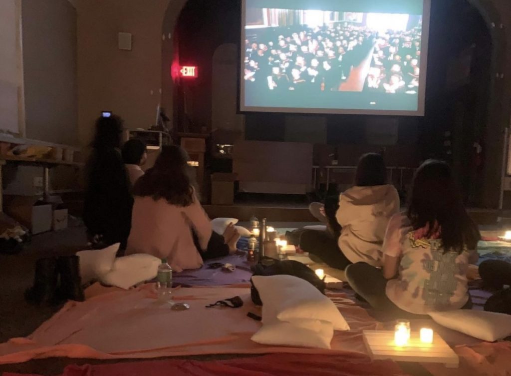Members of HSOC sit on blankets as they watch Legally Blonde on a projector screen. There are small, square, wooden planks with LED candles in between the blankets.