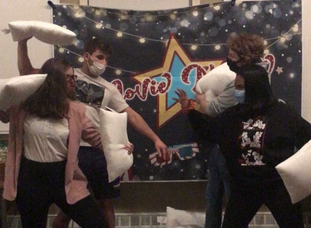Eli and Bella stand on the left, and Amiyah and Director Marques are on the right, all readying pillows. They stand in front of a tapestry that says "Movie Night" in a lavish, red, cursive font with a yellow and blue star behind it. The tapestry also has string lights and 3-D movie glasses on it.