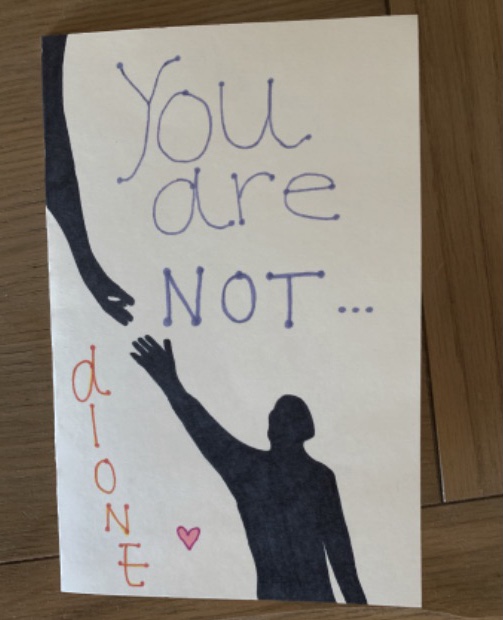 A drawing done with marker. A black hand reaches down from the left corner to a black figure holding out its hand on the bottom of the paper. On the top in purple marker, it says, "You are not..." and the word "alone" with a heart next to it is written on the bottom left.