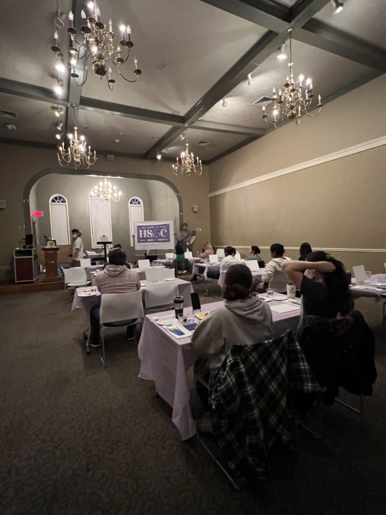 Students sit in pairs at tables with light purple tablecloths draped over them. There are brushes and art boards in front of each person. The HSOC logo  is on the whiteboard at the front of the Lyceum, and there is a Bob Ross cardboard cutout on the right of the whiteboard.