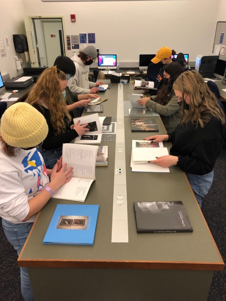 Students look through photobooks at a long rectangular table. There are seven students, and they wear mostly black shirts and jeans. The photobooks have a mix of text and colorful photos.