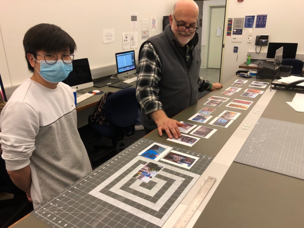 A student wearing a blue face mask, glasses, and a white shirt stands on the left and looks at the camera. The professor wears glasses and a sweater vest over a black and white plaid shirt. They're looking at many photos laid out over a rectangular table.