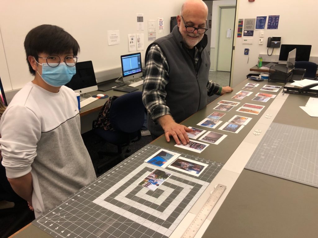 A student wearing a blue face mask, glasses, and a white shirt stands on the left and looks at the camera. The professor wears glasses and a sweater vest over a black and white plaid shirt. They&#039;re looking at many photos laid out over a rectangular table.