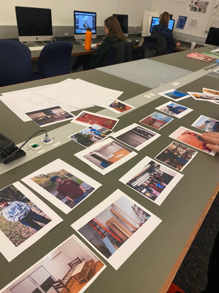 Many photos of various people and furniture are laid out on a rectangular table. Two students sit at computers in the background and edit photos.