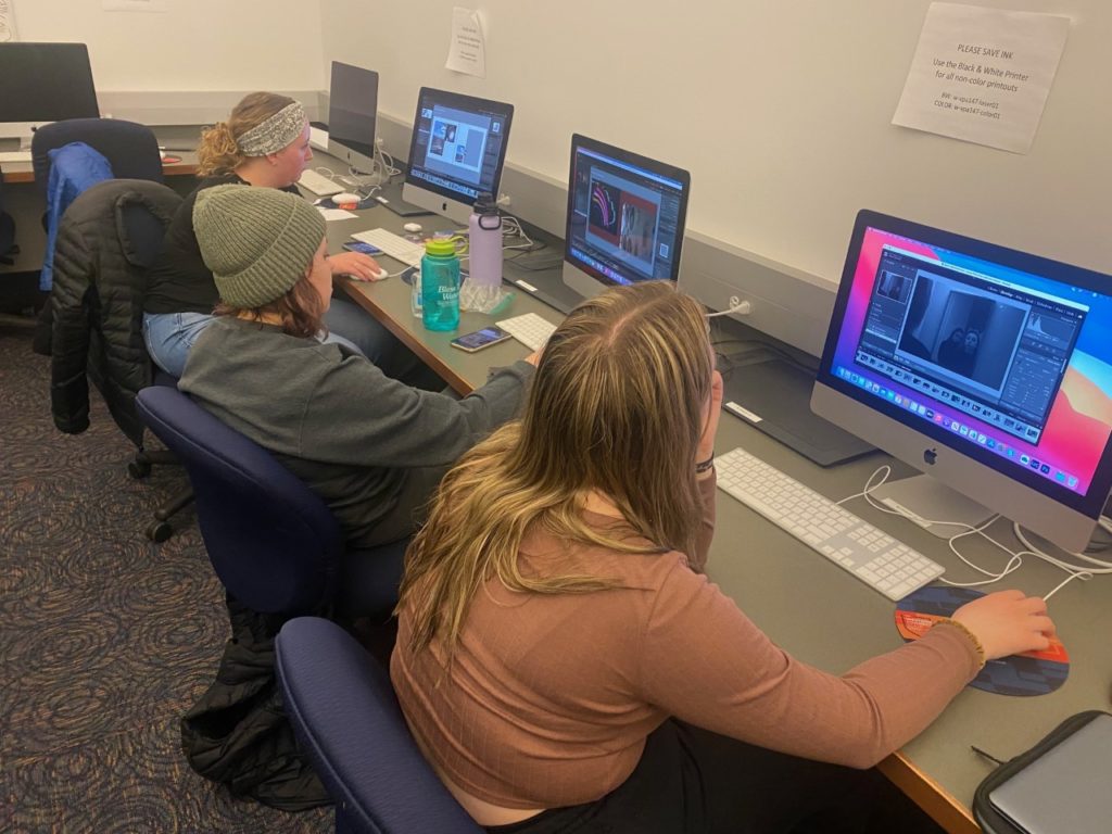 Three students sit at Macs and edit their photos. The student in the back wears a stretchy gray headband, a black shirt, and jeans. The student in the middle wears a sage green beanie and gray sweats. The student on the right wears a dark pink shirt and black leggings.