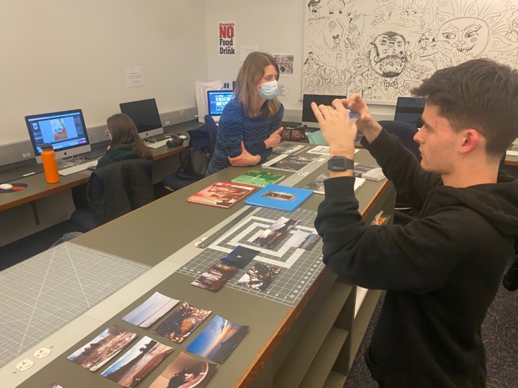 A student sits at a Mac in the background and edits a photo. The professor wears a blue sweater and blue face mask, and she leans on the rectangular table in the middle. A student stands in the foreground taking pictures of the photos arranged on the table. He is wearing a black sweatshirt.
