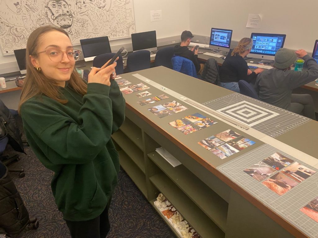 A student stands in front of the table with their phone in their hands, about to take a picture of the photos arranged on the table. They are wearing a dark green sweater and black leggings. Students sit in the background editing photos on the Macs.