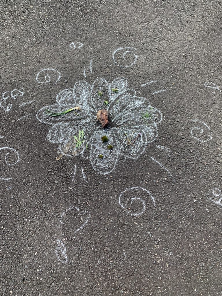A rock sits in the middle of a flower drawn with chalk. Moss and grass are strewn across the petals. Little spirals are around the flower.