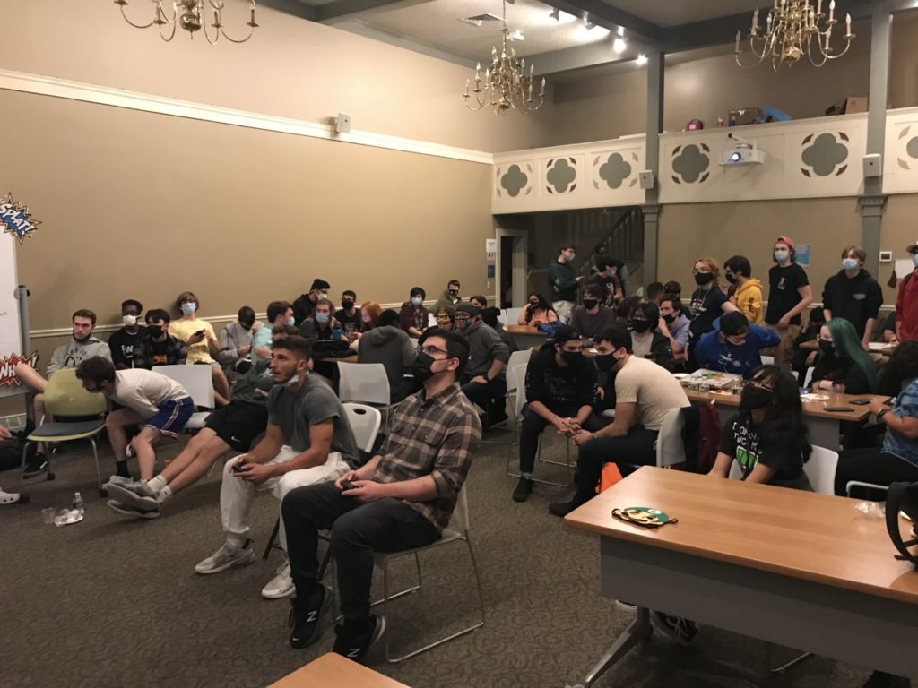 Many students sit and stand in the Honors House Lyceum as they watch two students in the front playing Super Smash Bros.