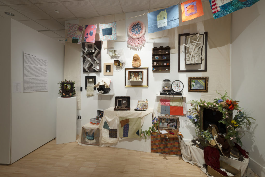 Many projects sit on display tables, on a back white wall, and pinned to a clothesline. Many of the projects have some kind of natural imagery in it and have plants attached to them. The projects on the clothesline are geometric and depict homes and/or comfortable spaces.