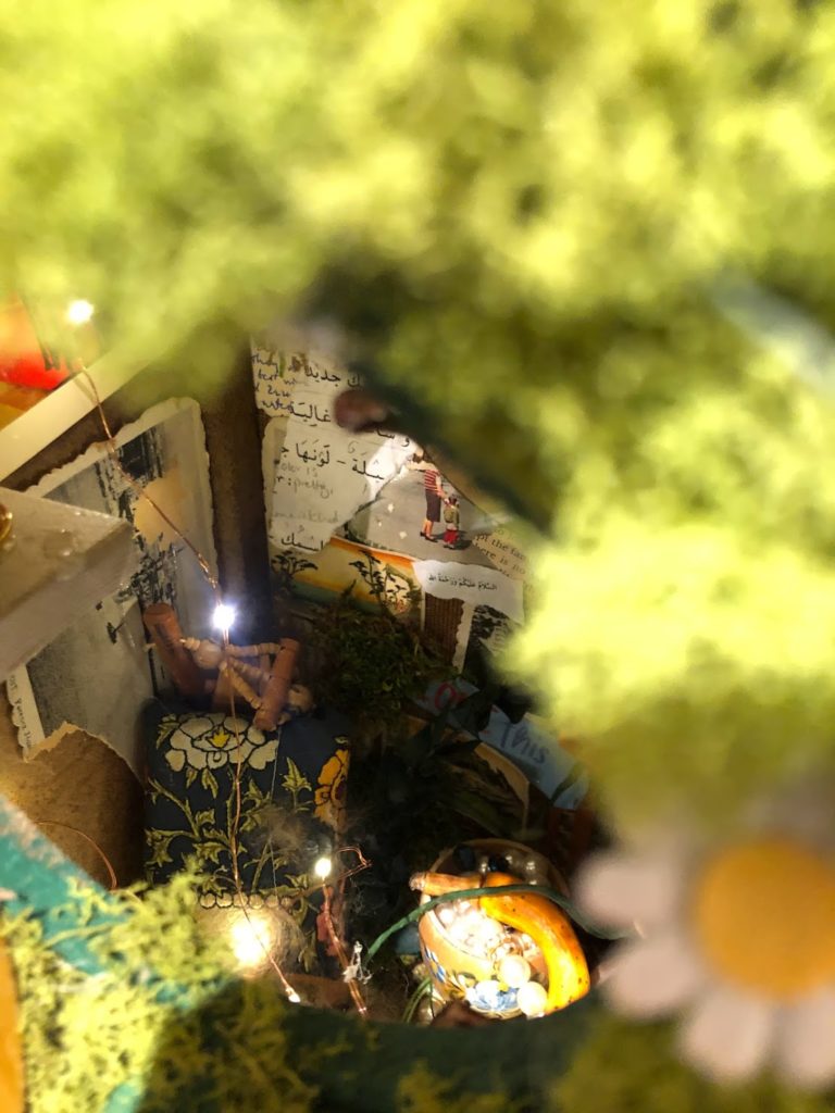 A view through the mossy roof of a tiny room. There are fairy lights and wooden chairs around the room. There's also a collage made of various writing and natural imagery on the back wall.