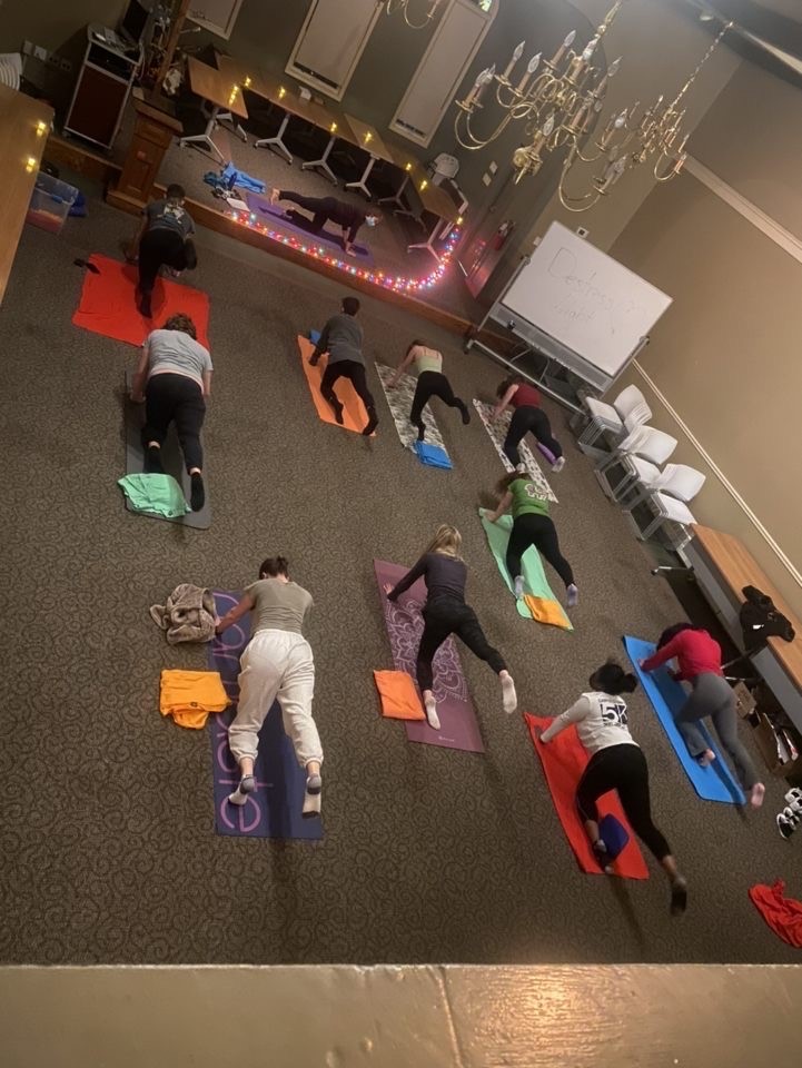Ten students do the bird dog yoga pose on top of blankets and yoga mats in the Honors Lyceum. Christel Autuori is at the front of the room on the raised floor on a yoga mat. Many tiny LED light candles form a half circle around the side of her yoga mat facing the students.