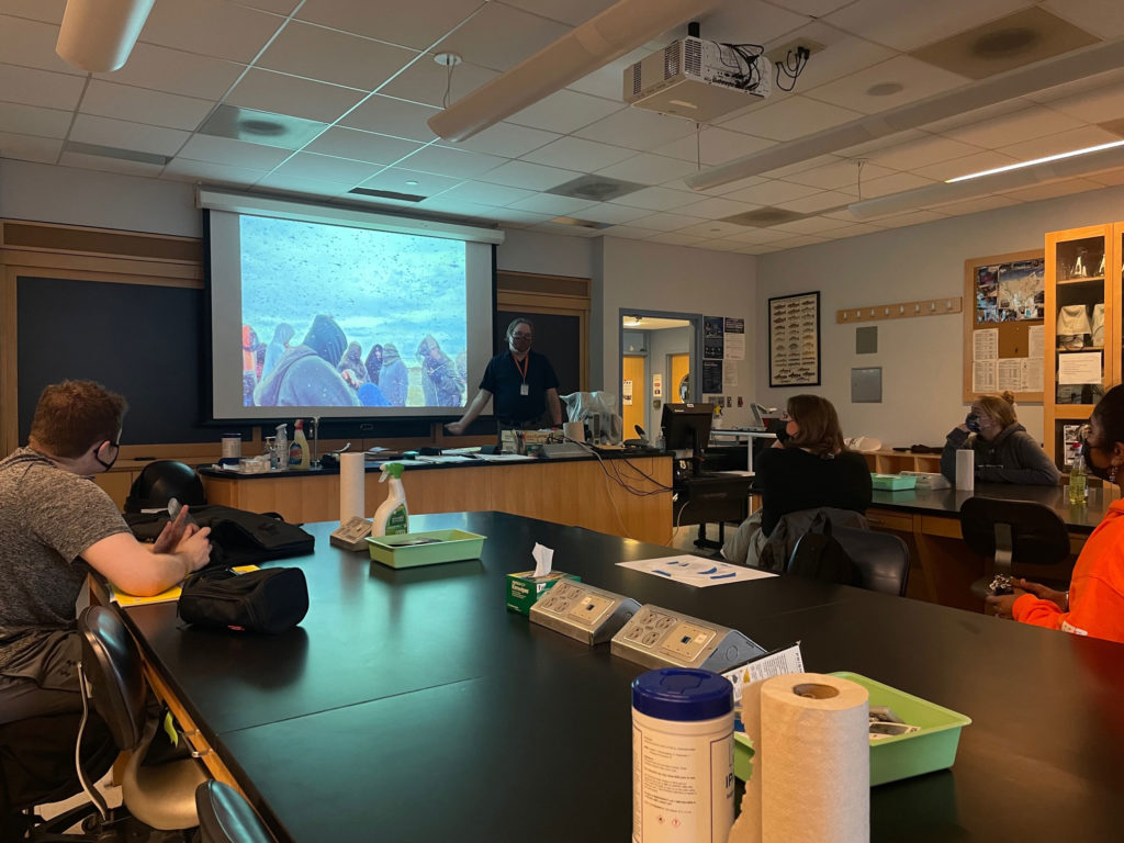 Biology professor Mitch Wagener gives a presentation about ecological and environmental biology. The students sit at long black tables and are all wearing face masks.