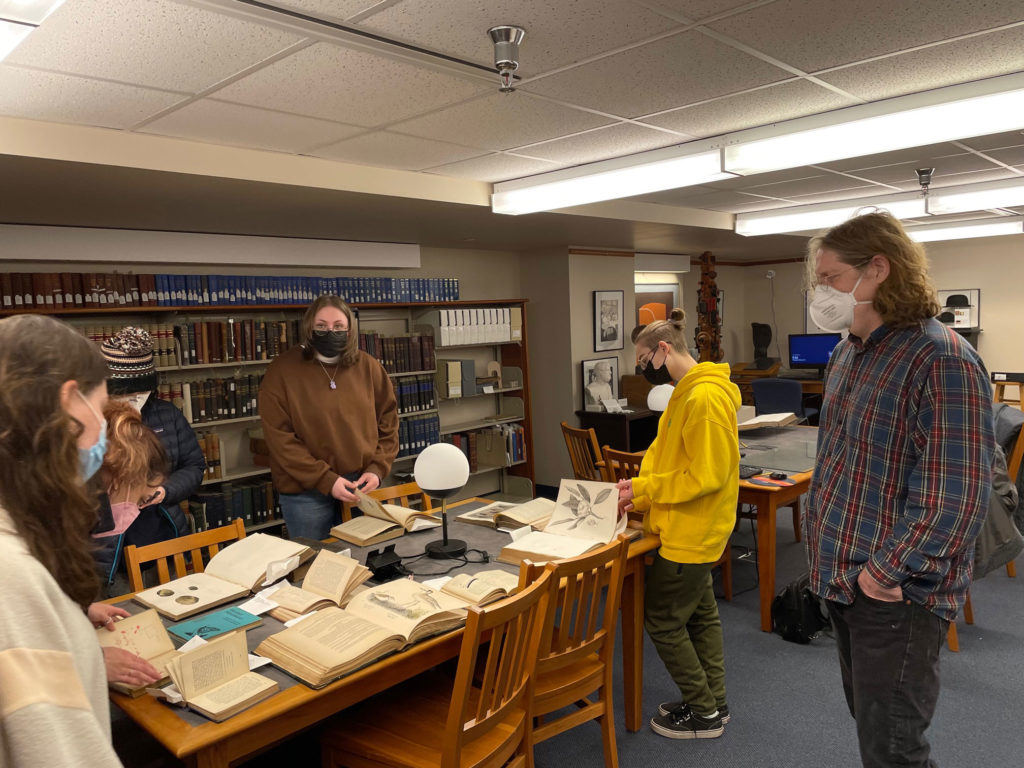 Students stand around a table with library archivist Brian Stevens. Many books are open on the table, and the students peruse the pages and look at drawings.