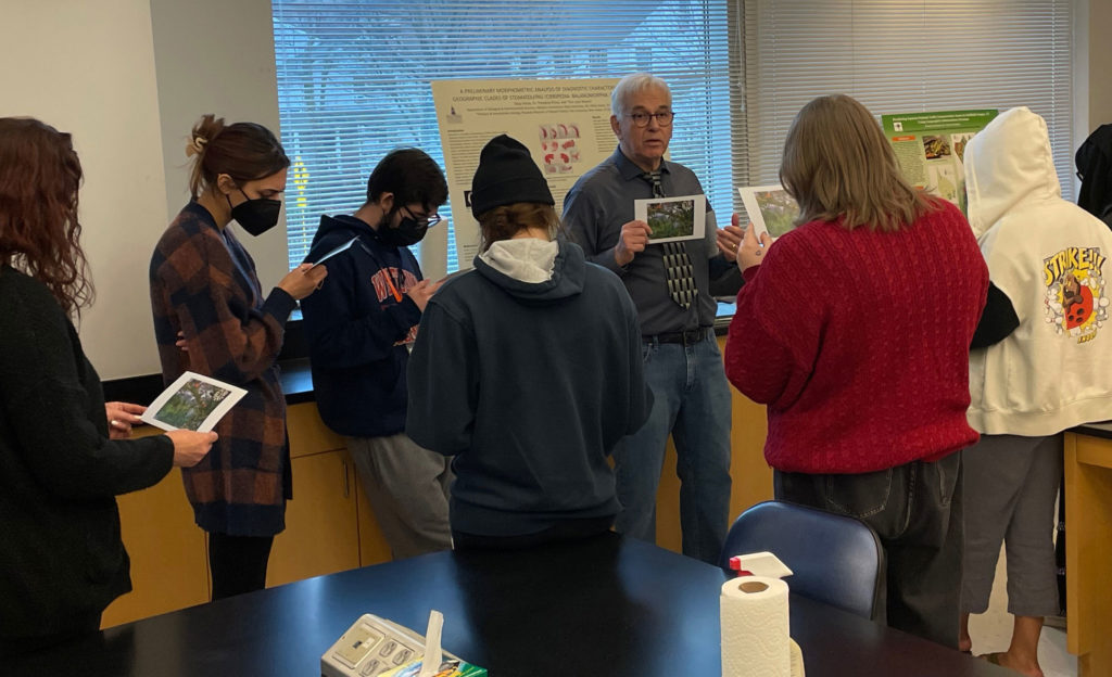 Michael Rothman is wearing a dark grey shirt, a black and grey tie, and jeans. Students wearing face masks look at copies of his work on cardstock.