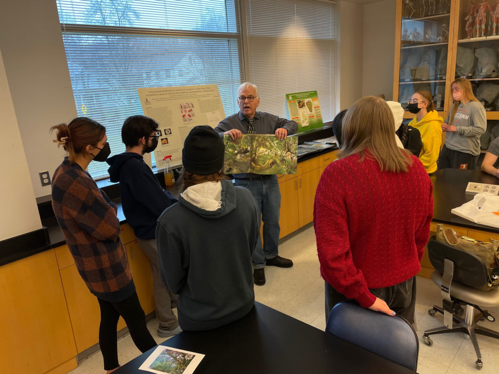 Students stand around Michael Rothman as he holds up a book with two hands. The open page depicts a very green illustration of a jungle.
