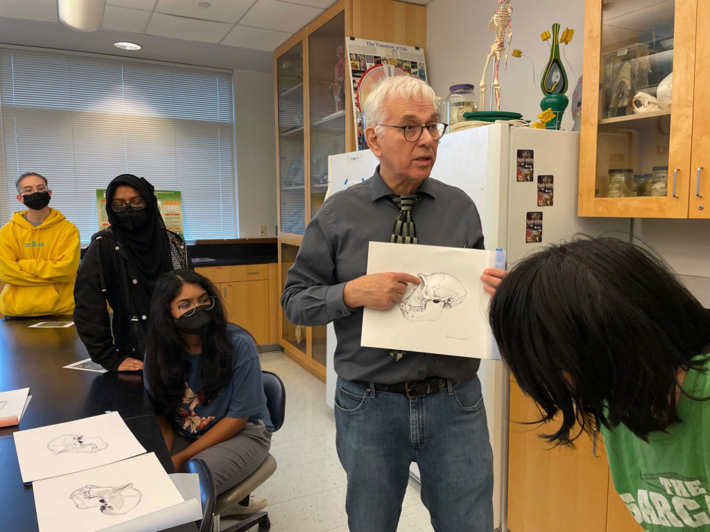 Michael Rothman is holding up a white piece of a paper with a monkey skull illustration printed on it. Students sitting at the surrounding lab tables look at him and the copies of the illustration on the tables.