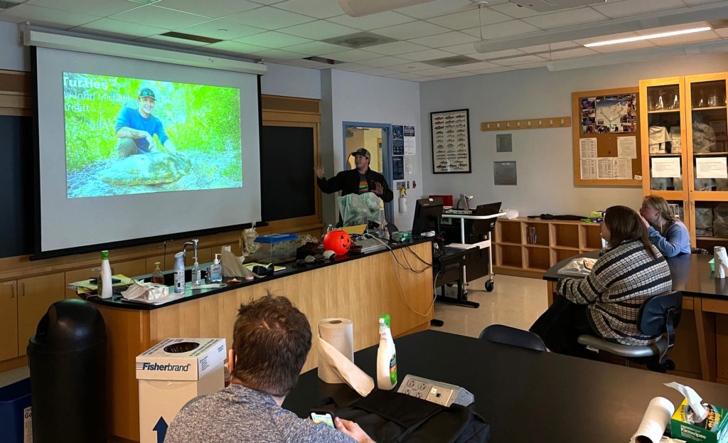 Arnett is gesturing to the presentation slide, which says in white font, "Turtles by John Michael Arnett." The background of the slide is a picture of Arnett with a large turtle shell.