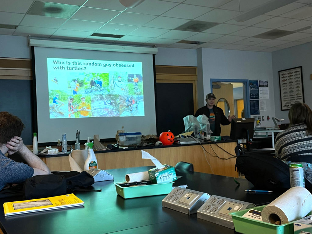 Arnett is giving a presentation to students about turtles. Behind them is a presentation slide that says "Who is this random guy obsessed with turtles?" in bold black font at the top. The rest of the slide is covered in photos of Arnett working with turtles.