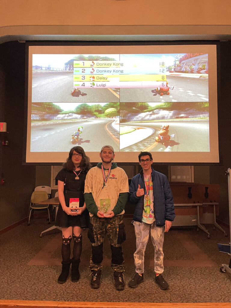 Three students stand in front of a projector screen in the Kathwari Honors House Lyceum. The screen shows the end of a Mario Kart game with four different windows, each one showing a different player. The students are wearing tiny medals, and the student in the center and the student on the left are holding up prizes they won.