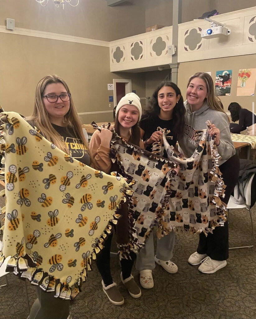 Four students stand in the Kathwari Honors House Lyceum holding up blankets. They are all dressed in sweaters and wearing comfortable shoes like Crocs, slippers, and sneakers. One blanket being held up has many bees on it, and the other has dogs of various colors. The blankets were made in a collaboration between the Honors Students of Compassion and Rotaract Club.