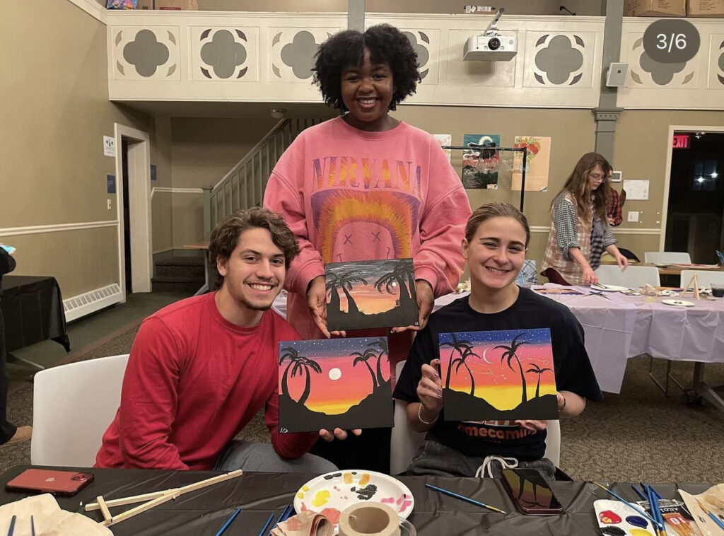 Three students hold up their paintings of sunsets at the HSOC Paint & Sip event in the Kathwari Honors House Lyceum. Each painting has palm trees and sand dunes painted in black. One student is standing behind the other two students who are sitting in chairs. In front of them is a table with paints, brushes, mini easels, and other art supplies.