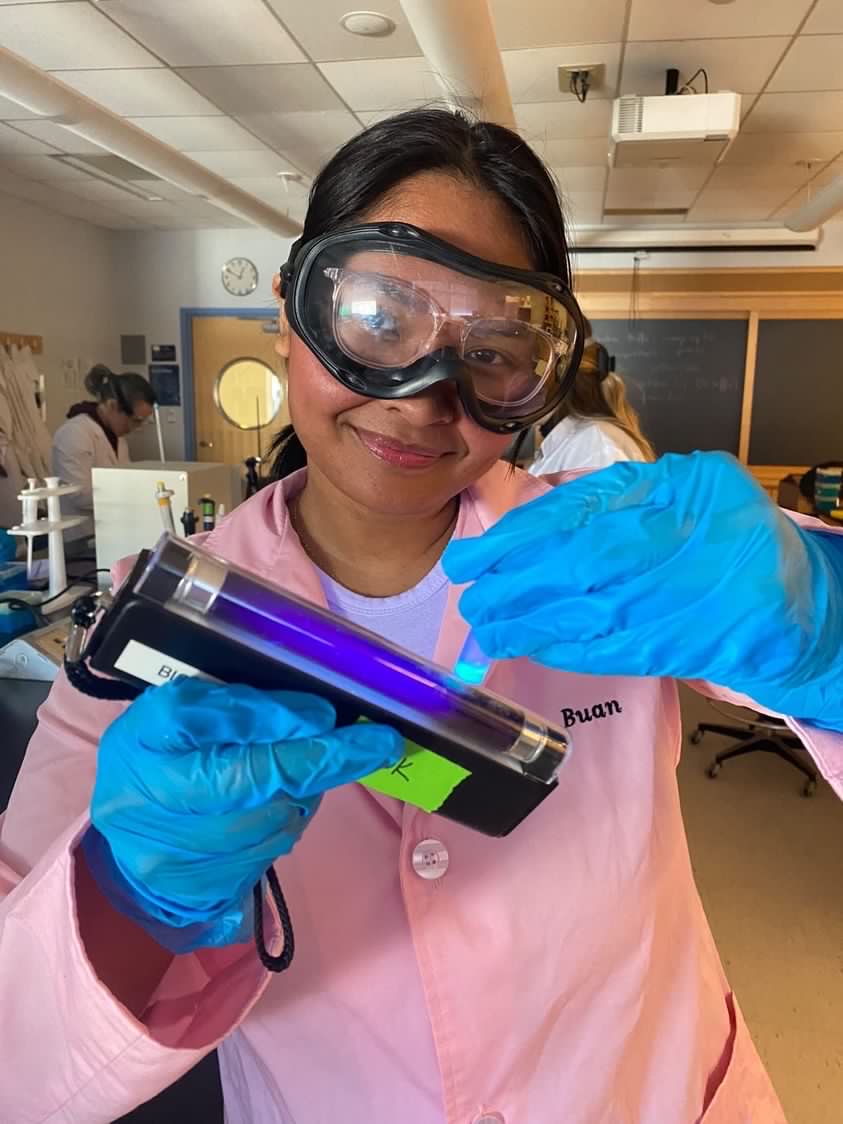 Amiyah is holding up a fluorescent light to a small tube with a small amount of glowing green liquid in it. Amiyah has medium brown skin and black hair. She is wearing large black goggles over a pair of white-rimmed glasses. She is also wearing a pink lab coat and blue gloves.