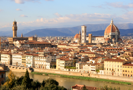 A helicopter view of the city of Florence, illuminated by a setting sun, with a river below and a mountainscape seen in the distance under a partly cloudy sky.