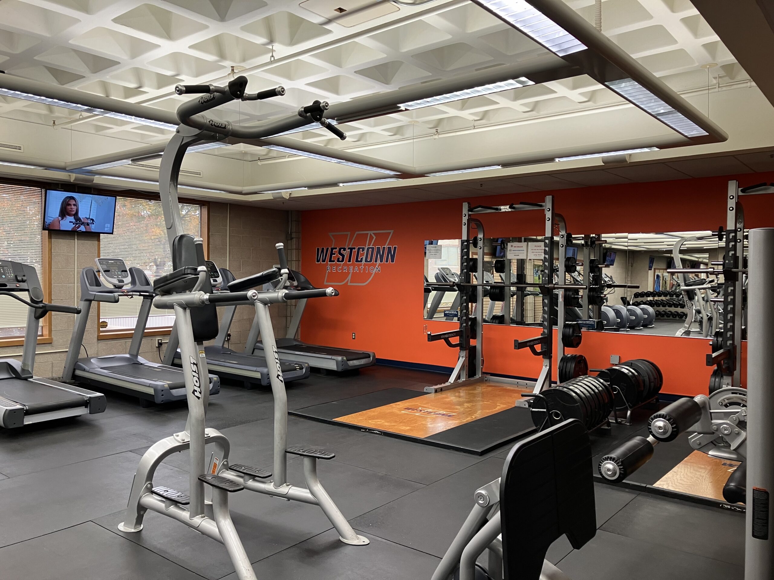 Fitness room showing weight and cardio equipment. WCSU branding on far wall, which is orange and has a mirror on it.